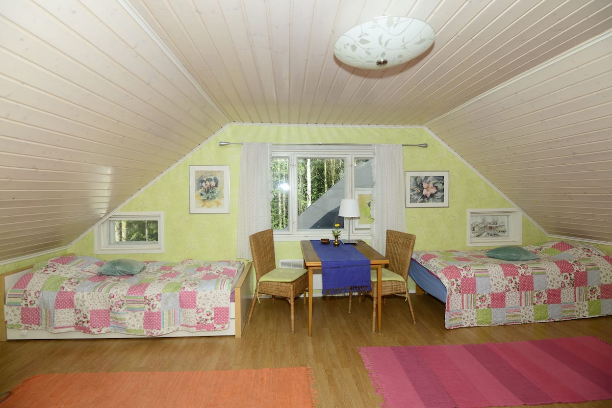 A cozy attic bedroom with two beds, a dining table, and a window with curtains.
