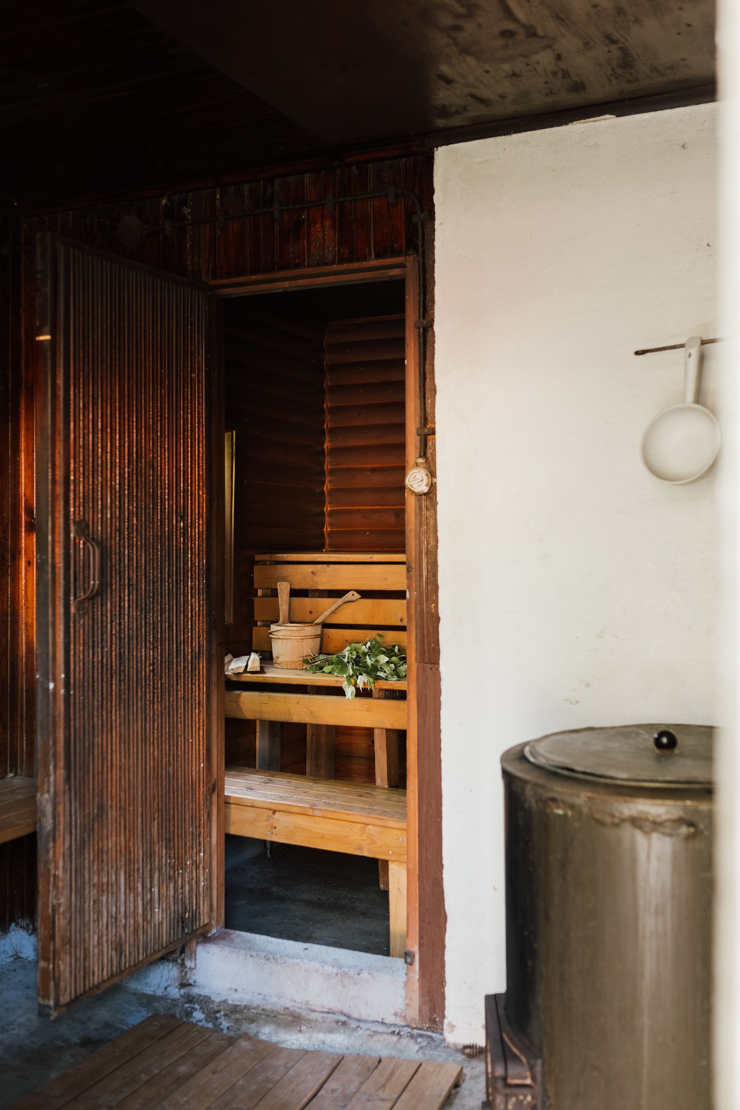 A rustic wooden door leading to a small room with a bench, a wooden bucket, and a potted plant.