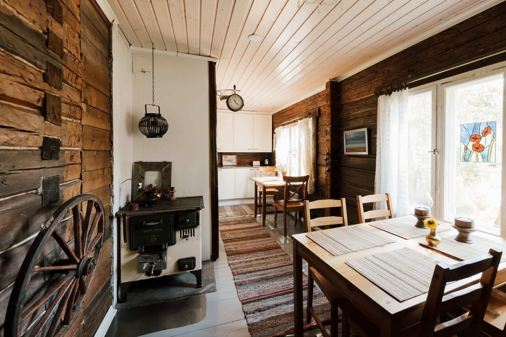 A rustic cabin interior with a dining area, wooden walls, a kitchen, and a large window with a stained glass view.