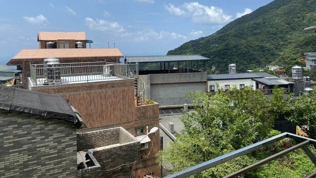 A multi-story building with a rooftop terrace, surrounded by greenery and a mountain in the background.