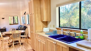 A kitchen with wooden cabinets, a blue sink, a window with a view of trees, and a dining area with chairs and a table.