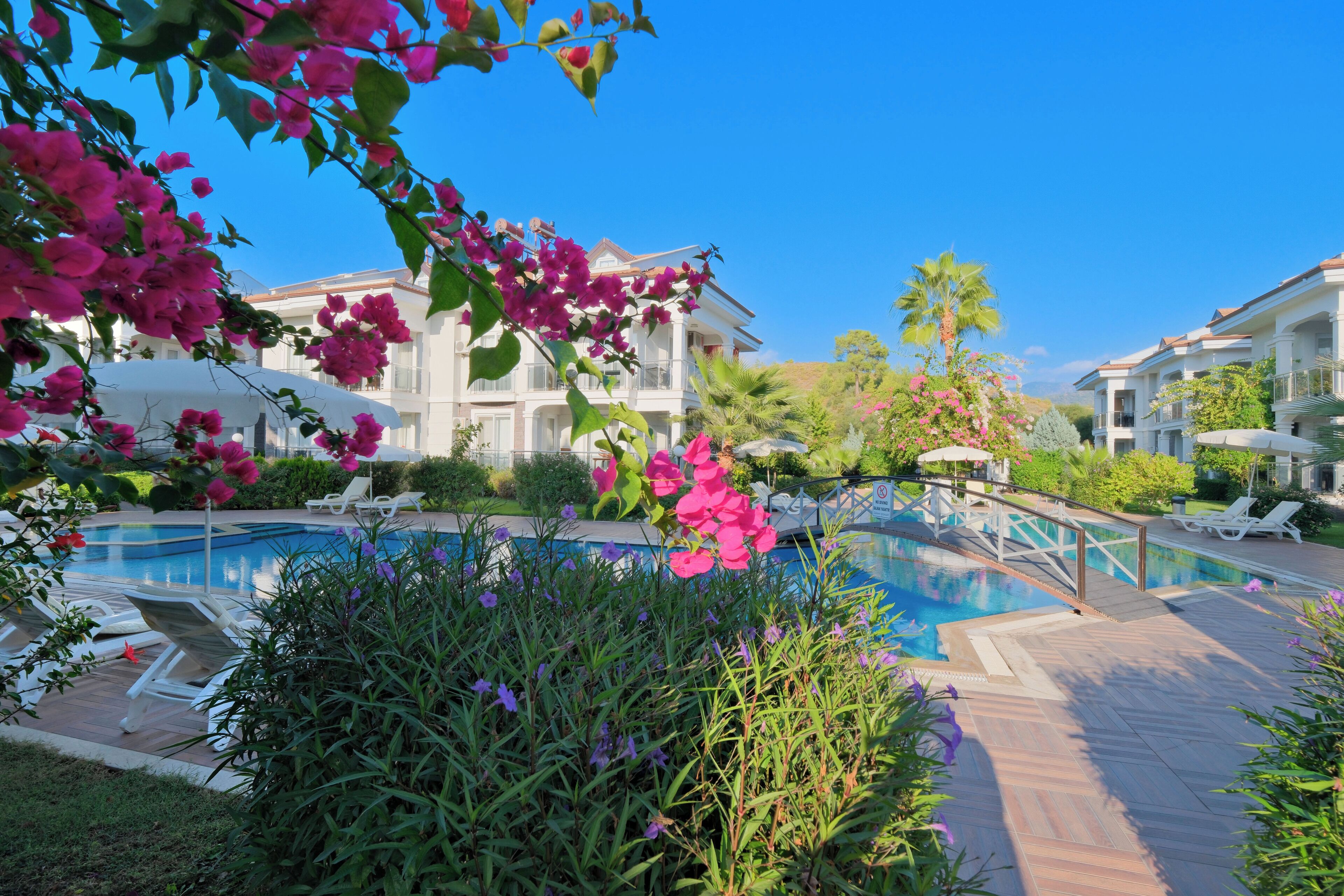 A pool area with lounge chairs, a wooden deck, and a building with balconies.