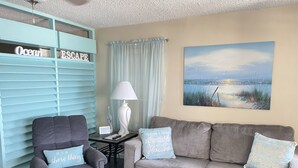 A living room with a grey sofa, a grey armchair, and a teal shelving unit with 'Ocean Escape' signage. There's a painting of a beach scene on the wall, a lamp on a side table, and a remote control on the sofa. - GULF VILLAGE CONDO LOCATED ON THE BEACH, DIRECT VIEW OF THE GULF, Sleeps 4. (Gulf Shores)