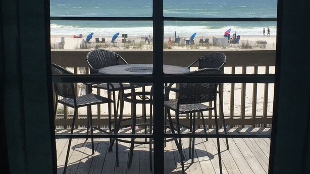 A deck with a table and chairs overlooking a beach with umbrellas and people. - GULF VILLAGE CONDO LOCATED ON THE BEACH, DIRECT VIEW OF THE GULF, Sleeps 4. (Gulf Shores)