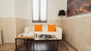 A modern living room with a white sofa, orange cushions, a glass coffee table, and a cityscape painting on the wall. - Hostal Live Barcelona (Barcelona)
