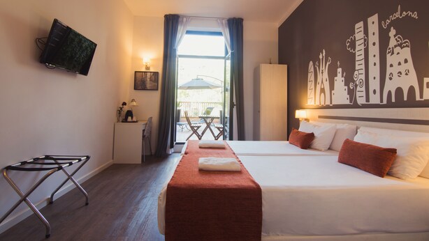 A hotel room with a bed, a TV mounted on the wall, a desk with a chair, and a view of the cityscape through the window. - Hostal Live Barcelona (Barcelona)