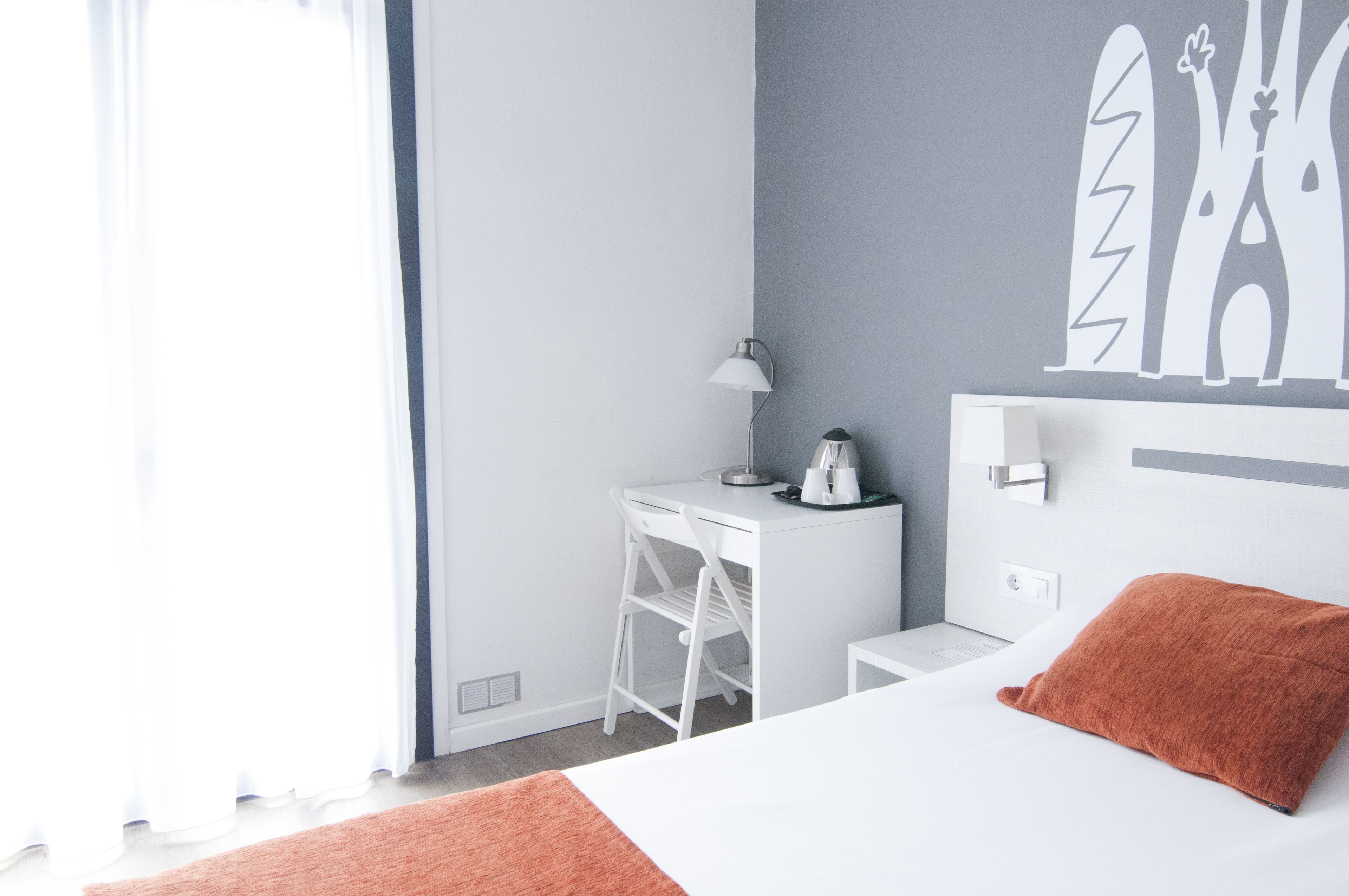Ein modernes Hotelzimmer mit weißem Schreibtisch, einer Lampe, einem Bett mit orangefarbenem Kopfkissen und einem Wandbild.