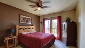 Una camera da letto con un letto, un comodino in legno con una lampada, una ventola a soffitto, una finestra con tende e un comò in legno.