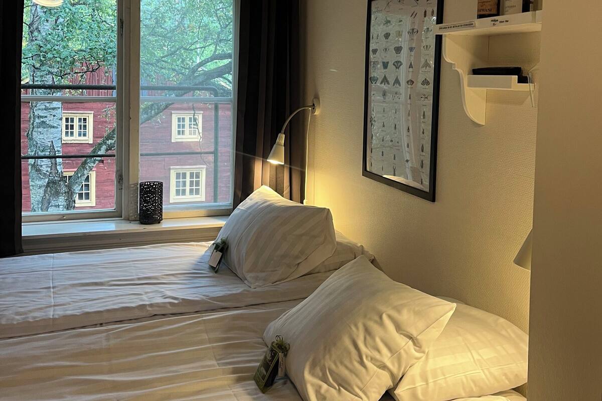 A hotel room with two beds, a window with a view of buildings, a wall-mounted shelf with bottles, and a framed picture.