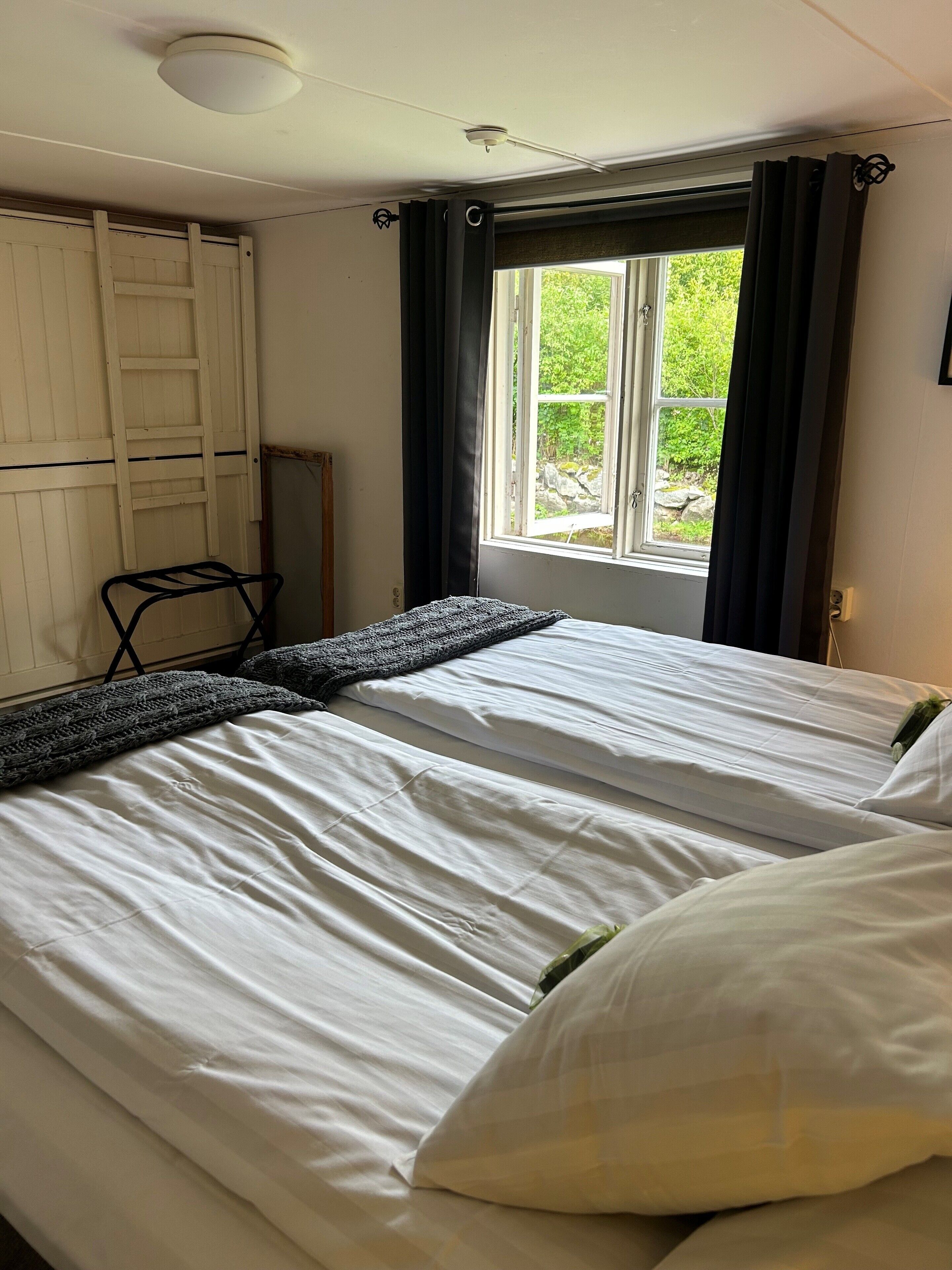 Un dormitorio con dos camas, una ventana con cortinas, una lámpara de techo blanca y una escalera de madera apoyada en la pared.