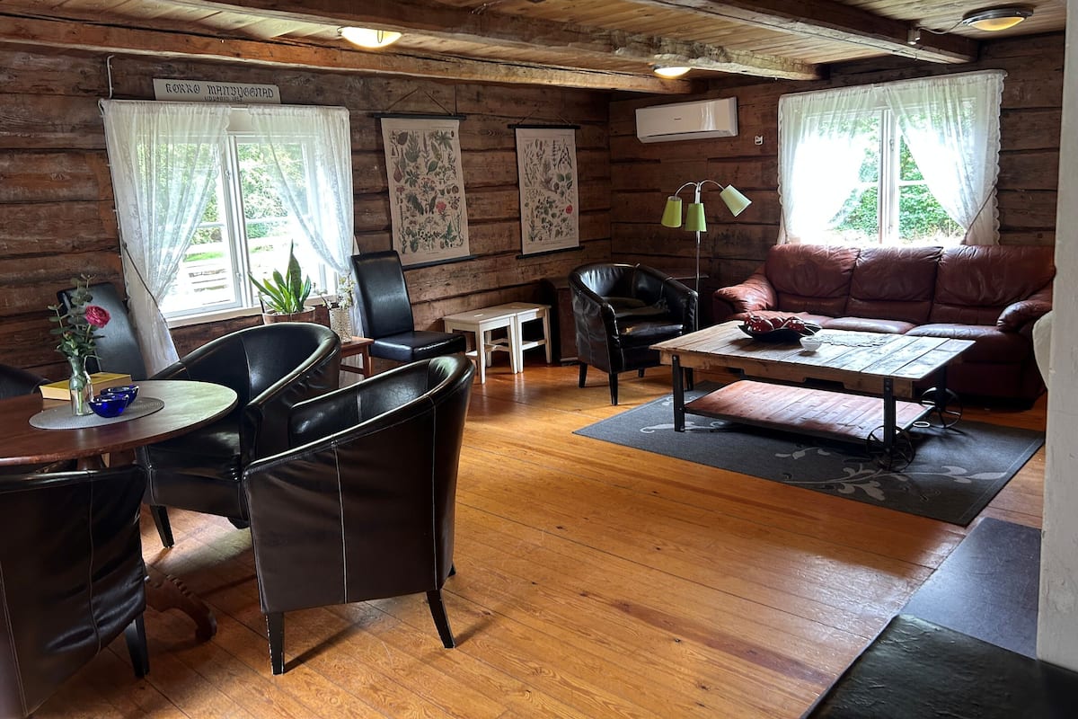 A cozy cabin interior with wooden walls, a leather sofa, a dining table with chairs, and a small kitchen area.