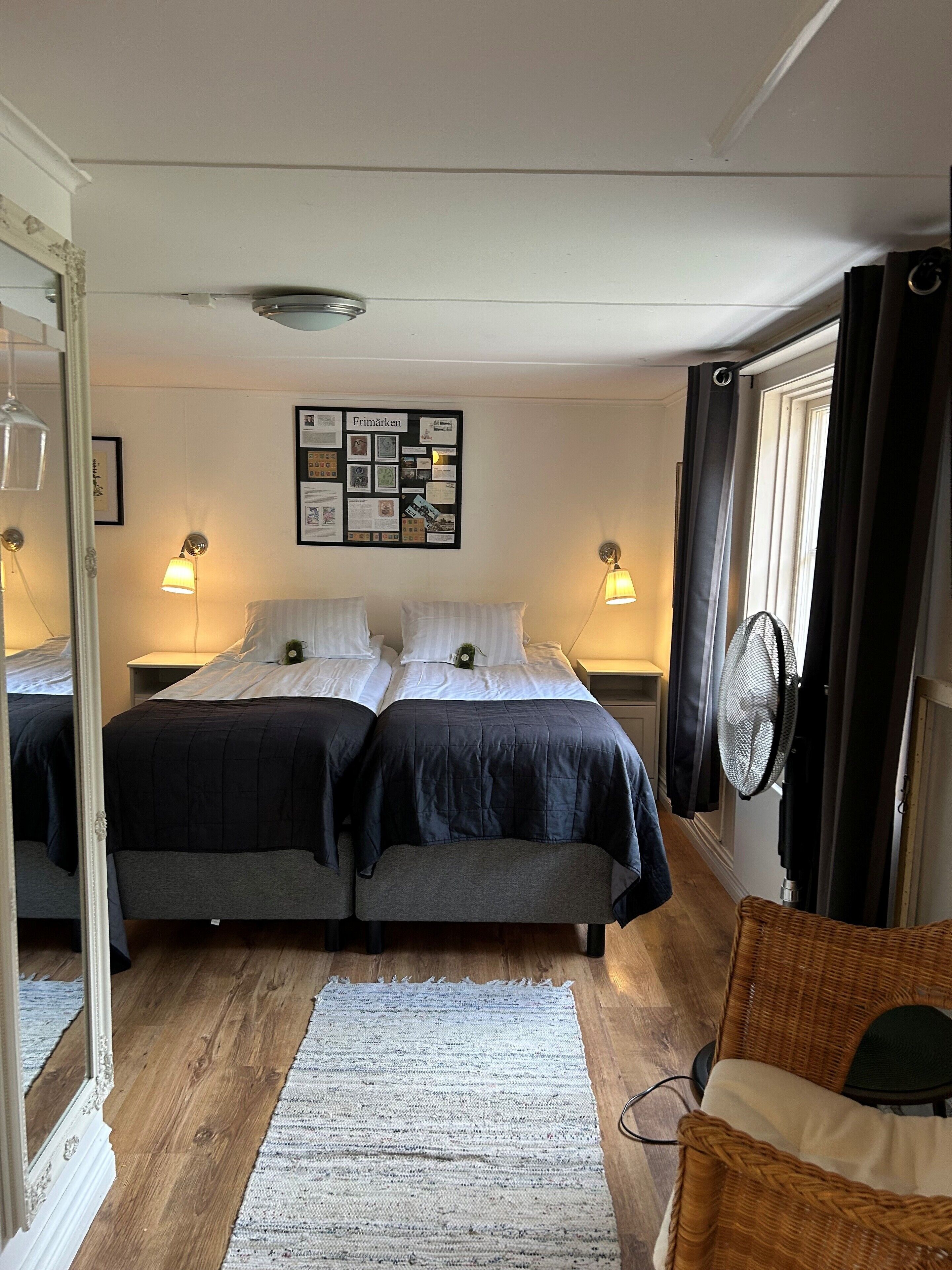 Ett sovrum med två sängar, en anslagstavla som hänger på väggen, sänglampor, en golvfläkt, en flätad stol och en spegel.