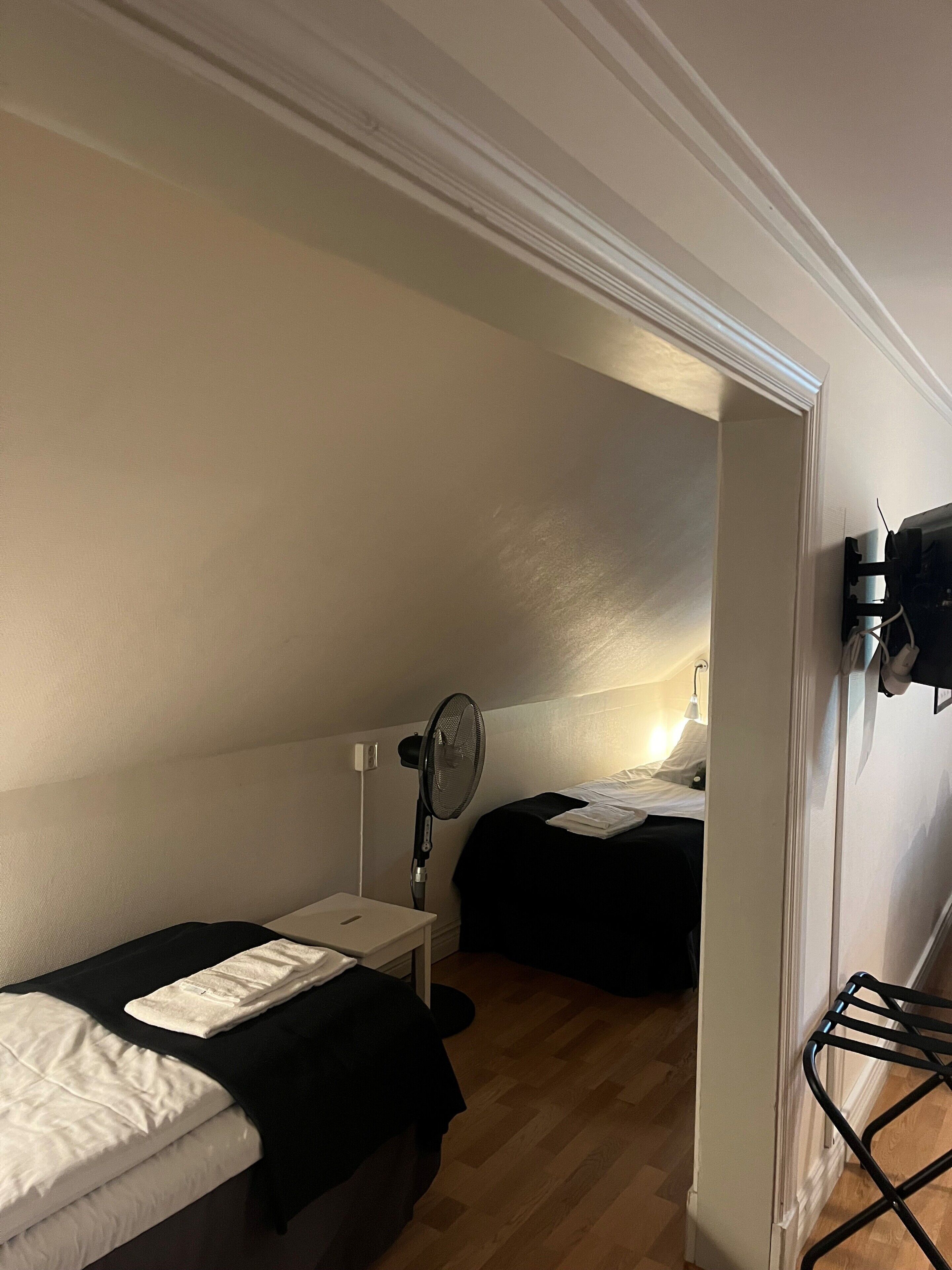 Ein kleines, schlichtes Zimmer mit einem Einzelbett, einem Nachttisch, einem Sessel und einem Standventilator.