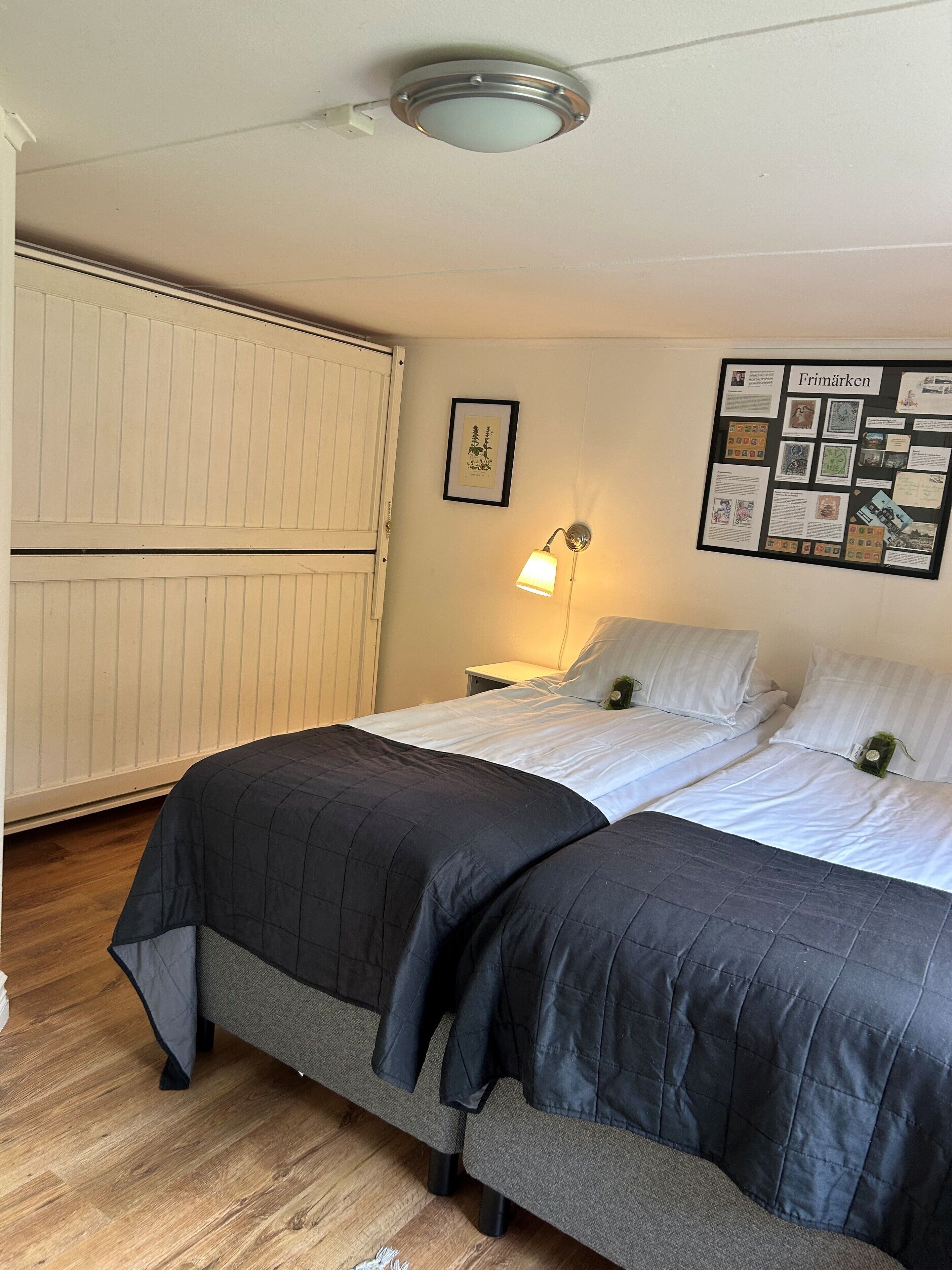 Une chambre avec deux lits, une table de chevet, une lampe fixée au mur et un tableau d’affichage où sont accrochés divers objets.
