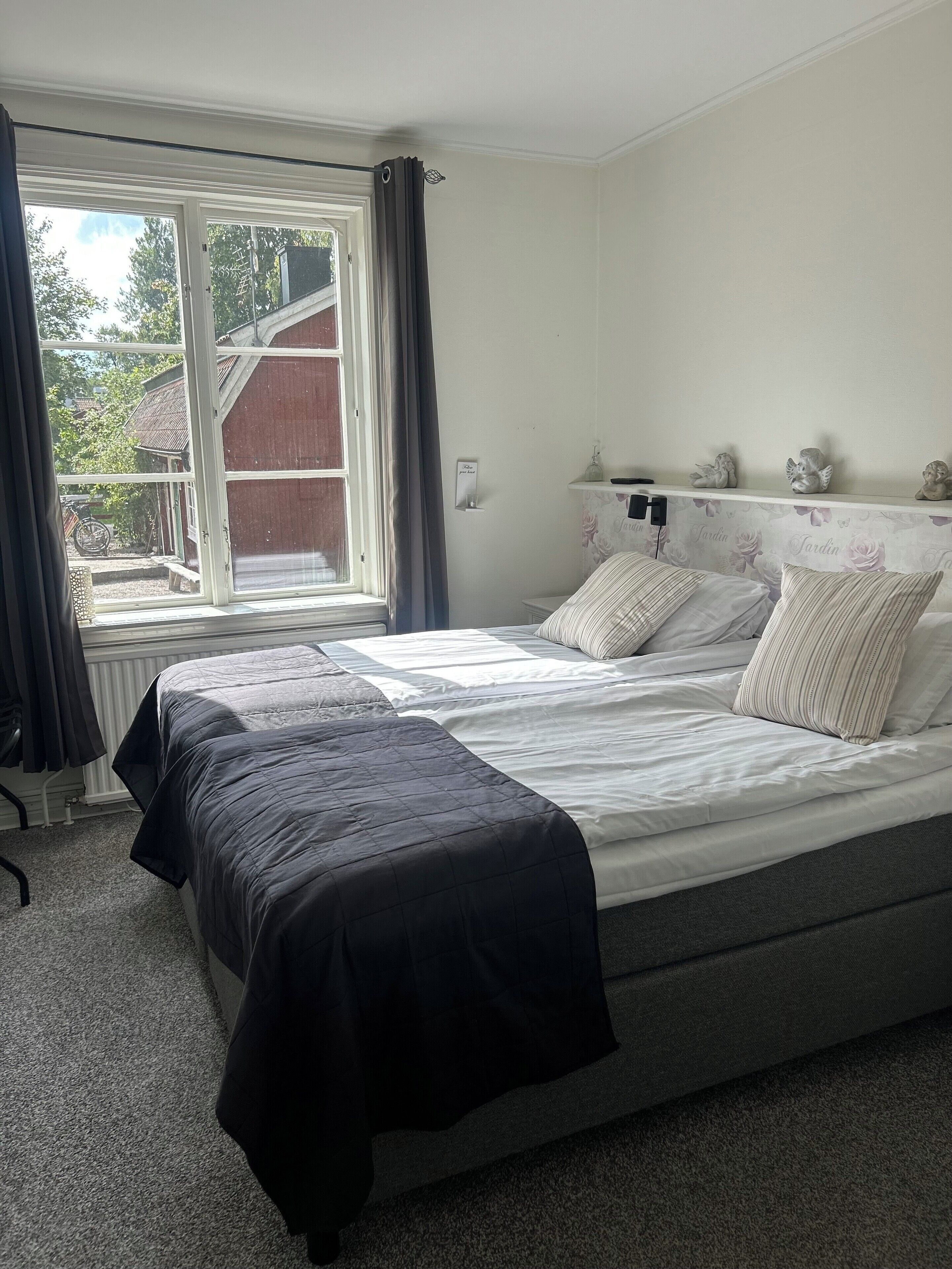 A bedroom with a large bed, a window with curtains, a radiator, and a small shelf with decorative items.