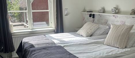 A bedroom with a large bed, a window with curtains, a radiator, and a small shelf with decorative items.