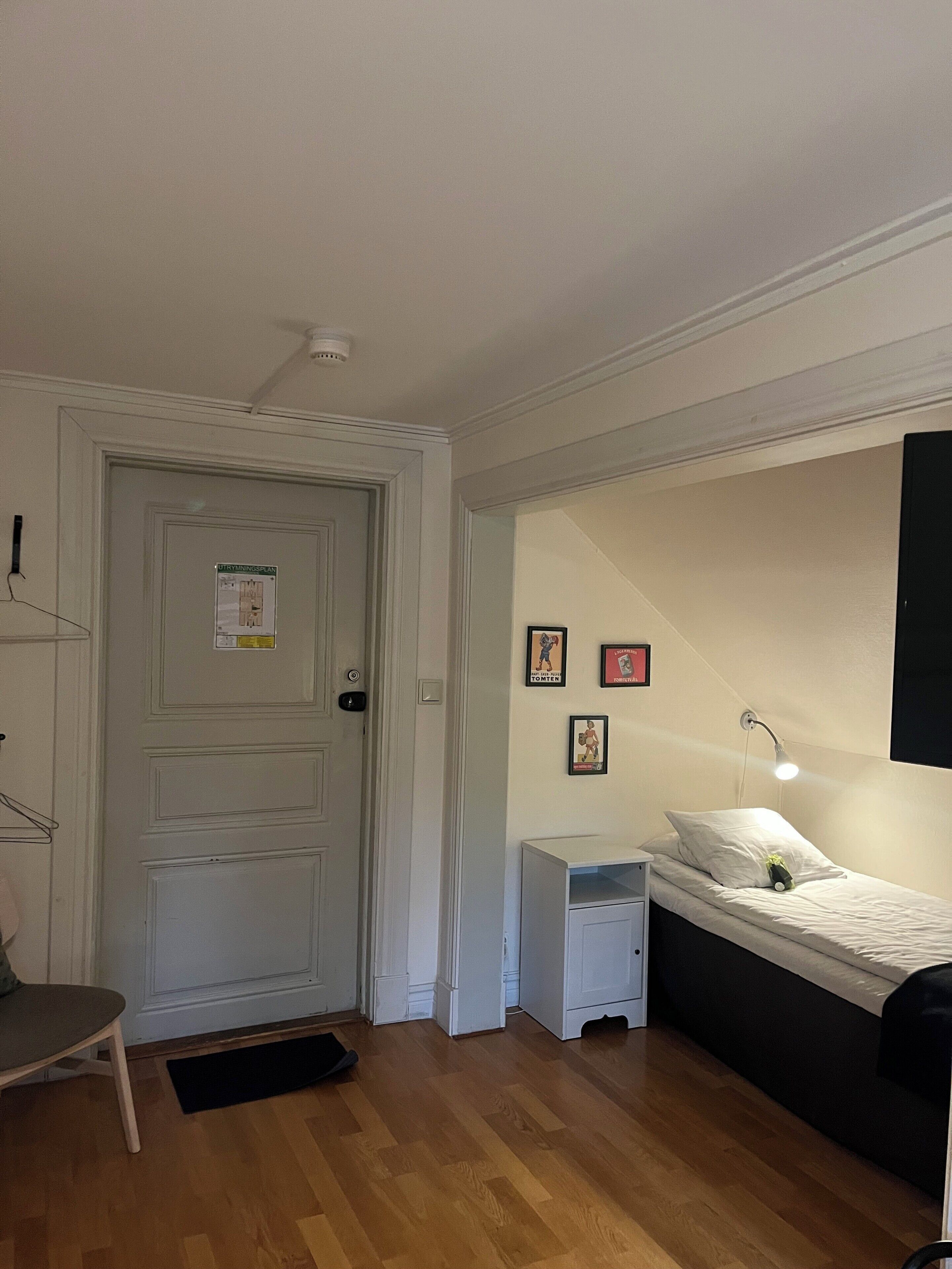 Quarto com cama, mesa de cabeceira, abajur e porta com pôster.
