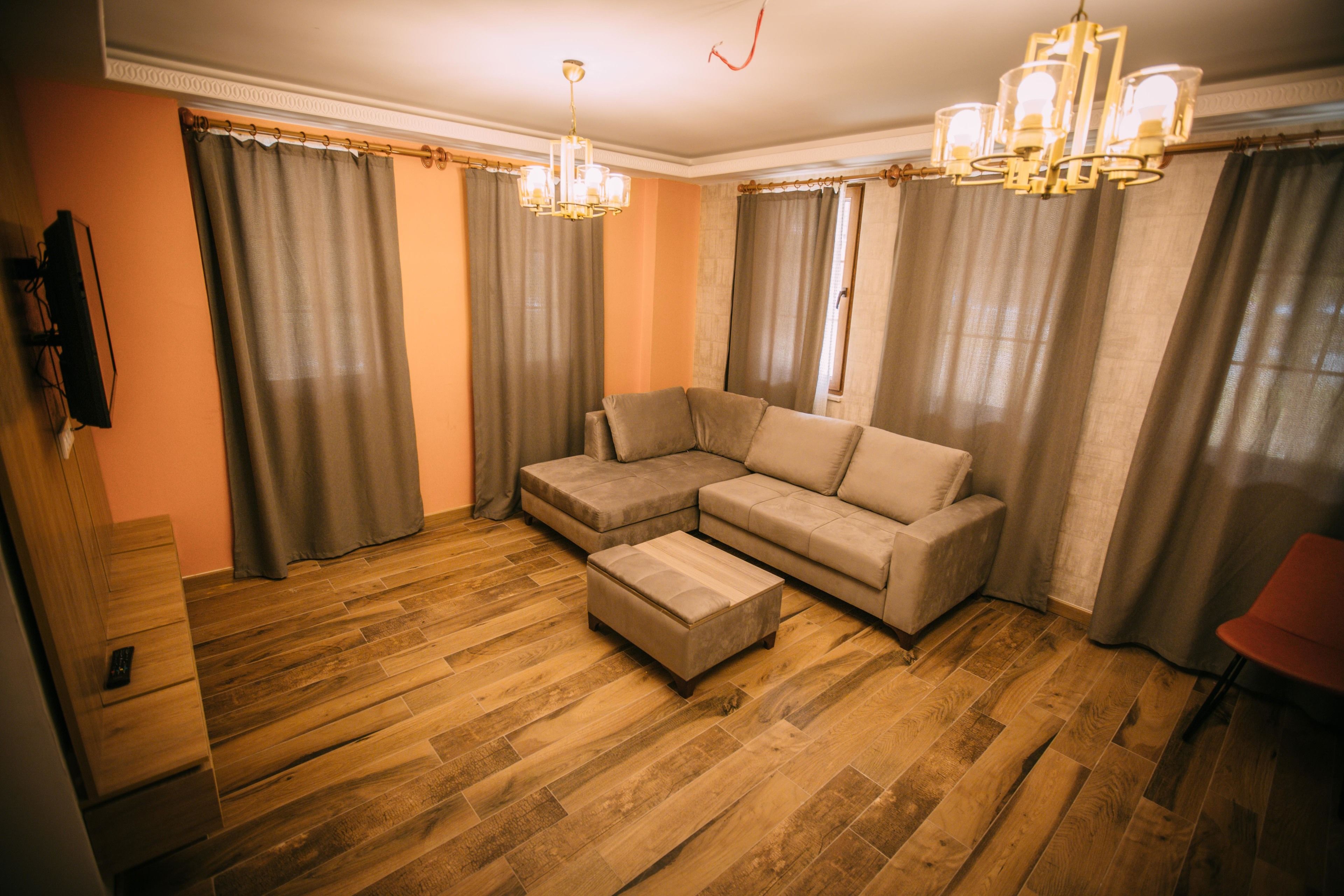 A living room with a sectional sofa, ottoman, wooden flooring, and a TV mounted on the wall.