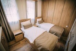 A compact bedroom with two beds, wooden walls, a small bedside table, and a window with curtains.
