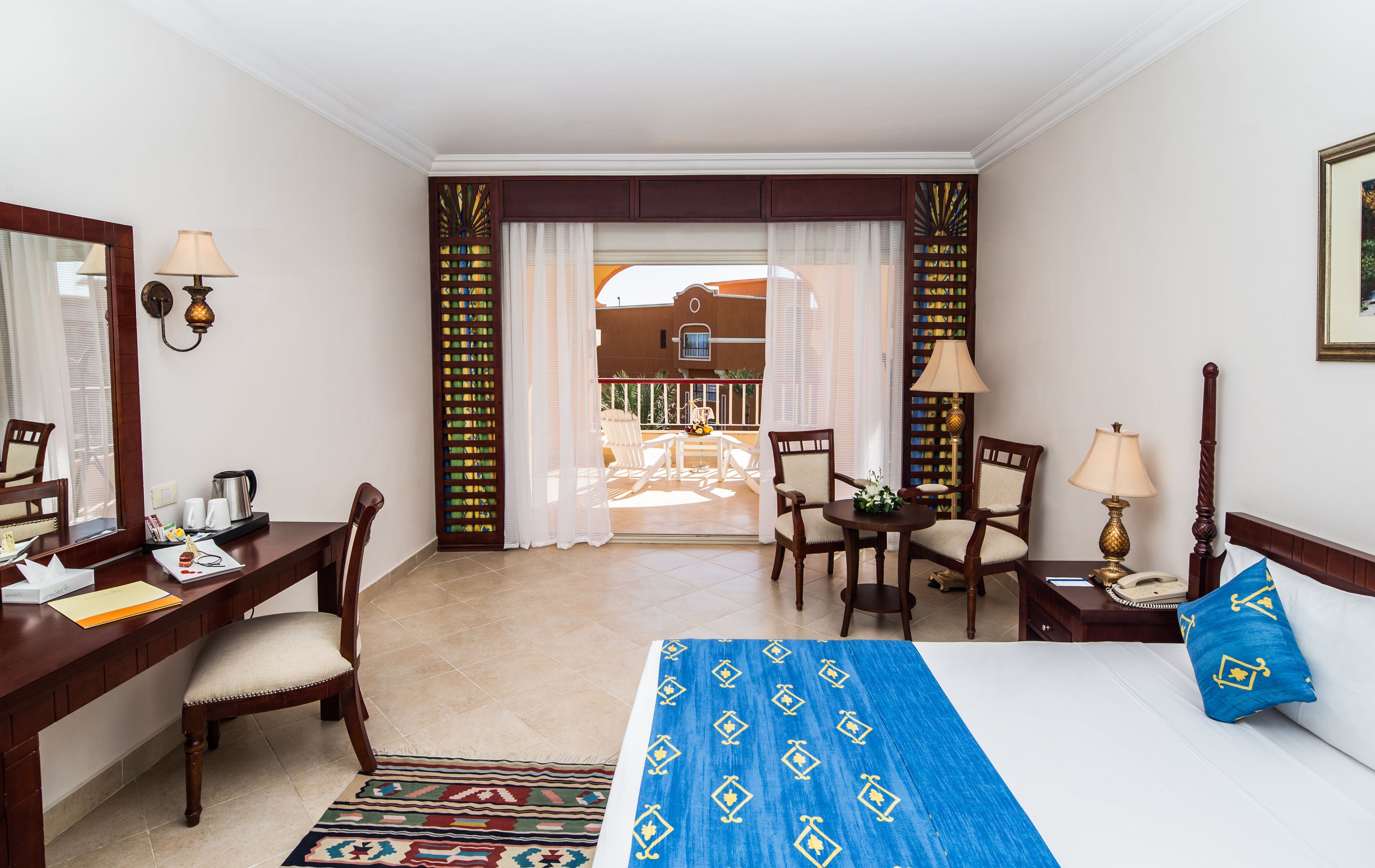 Habitación de hotel con cama, escritorio, dos sillas, una mesita, lámpara y vista al balcón exterior.