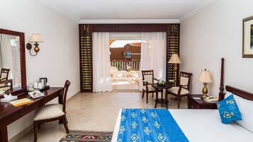 Habitación de hotel con cama, escritorio, dos sillas, una mesita, lámpara y vista al balcón exterior.