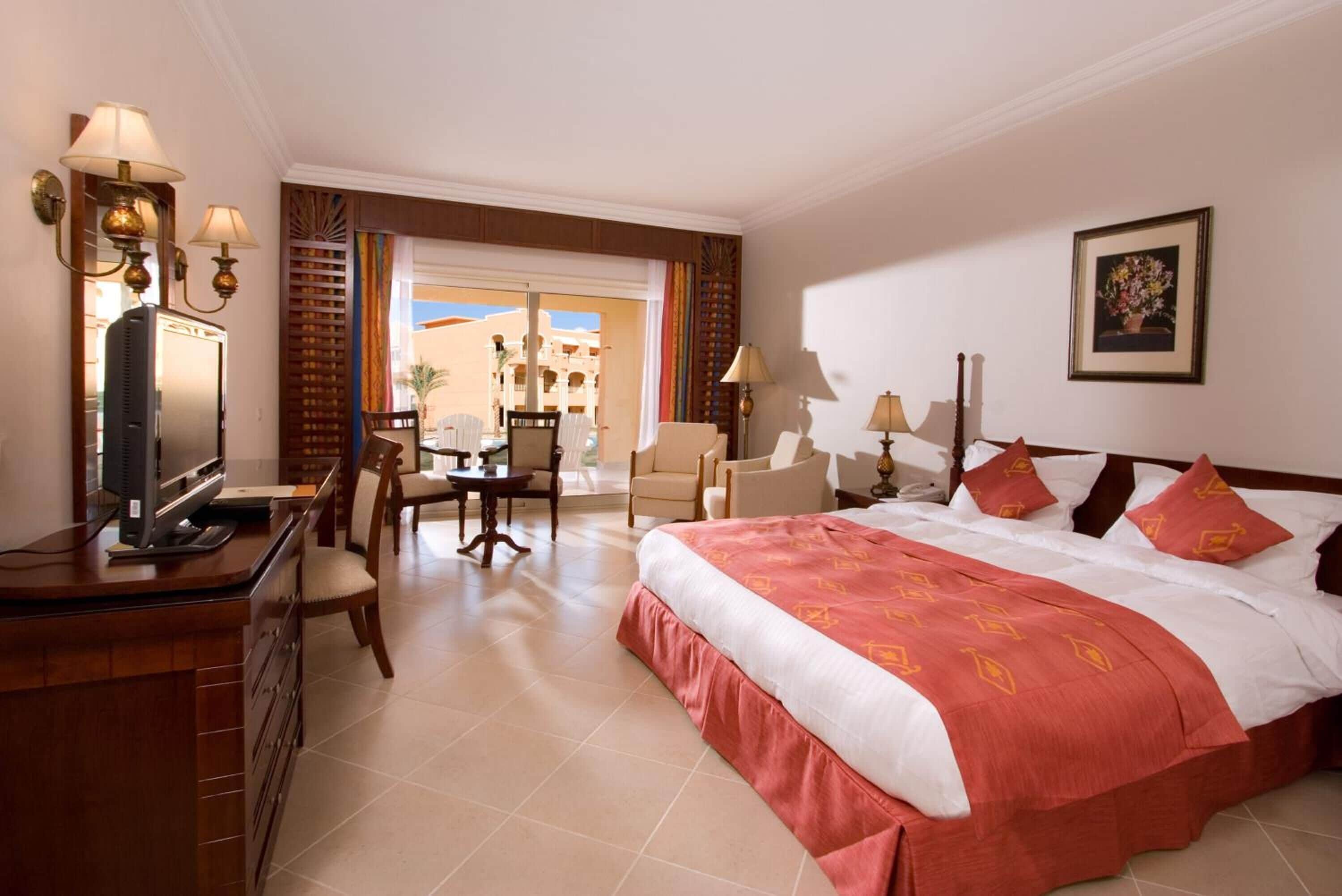 Habitación de hotel con una cama grande, un escritorio con televisor, zona de comedor con sillas y vistas al exterior.