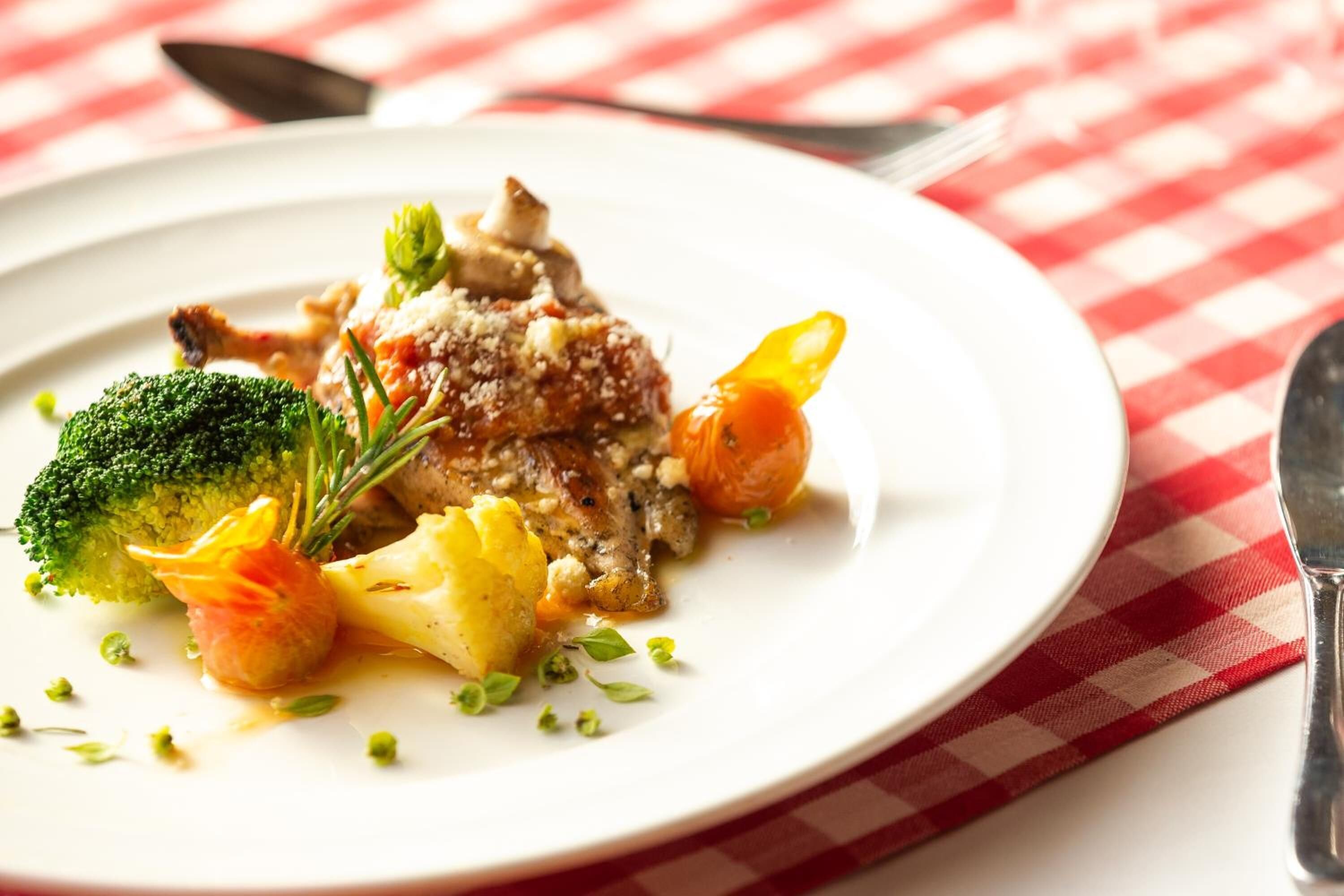 En tallerken mad med broccoli, en tomat og et stykke kød, serveret på en hvid tallerken med et rødt og hvidt ternet dug.