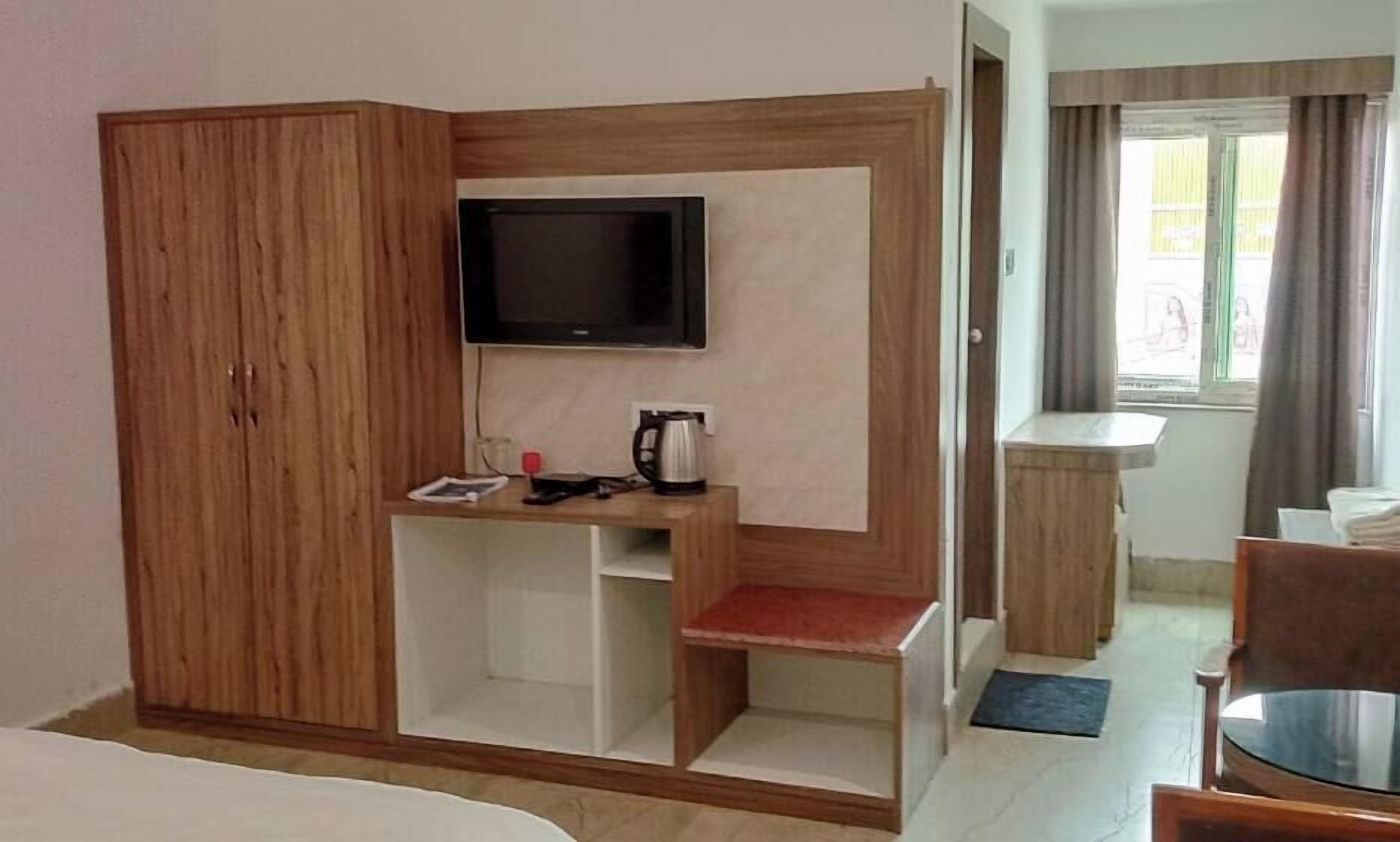 A hotel room with a wooden wardrobe, a TV mounted on the wall, a small table with a kettle, and a window with curtains.