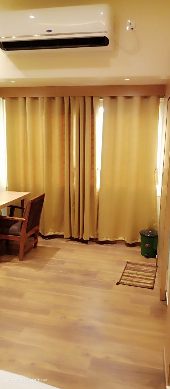 Una habitación con piso de madera, una mesa de comedor con sillas, un basurero verde y un ventanal con cortinas amarillas.