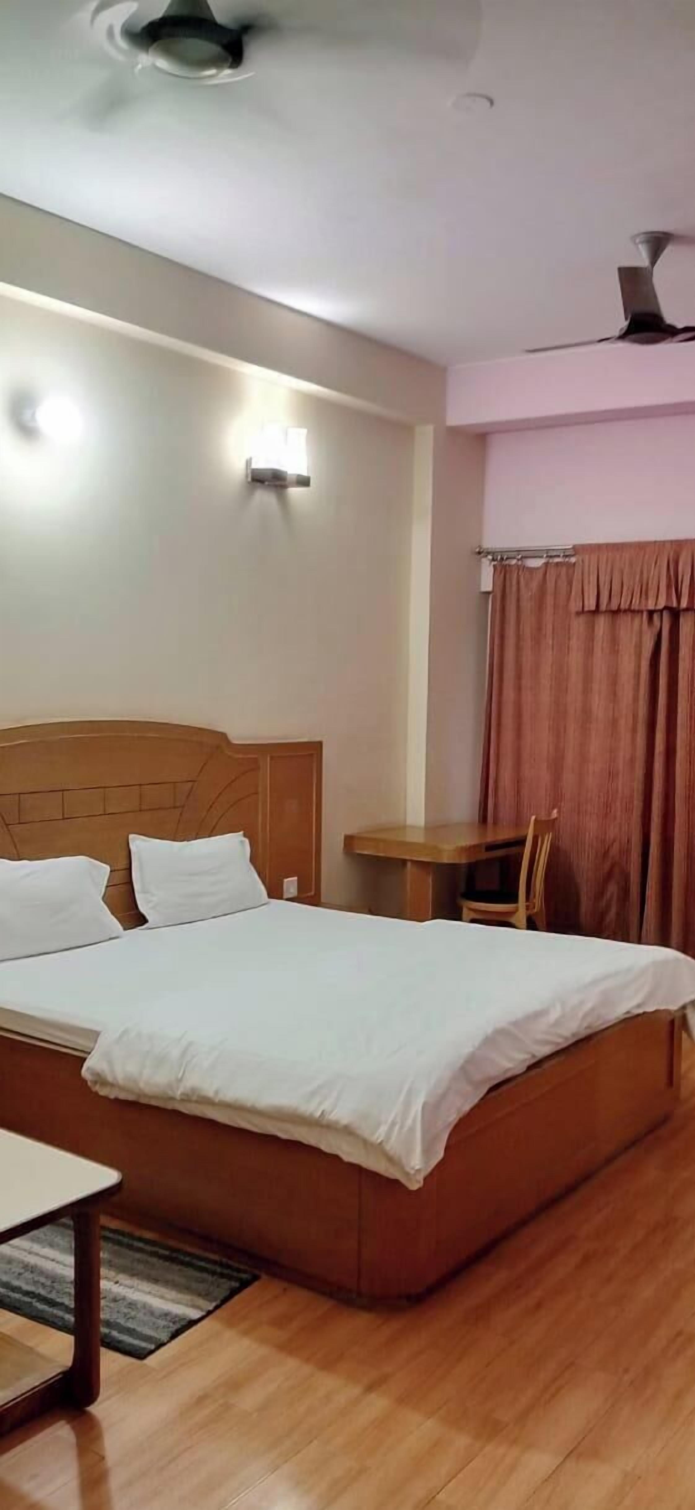 Habitación de hotel con cama, una mesa, una silla, ventilador de techo y luz empotrada en la pared.