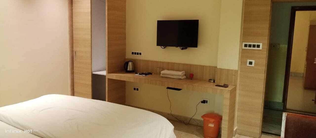 A hotel room with a bed, a TV mounted on the wall, a small table with a kettle, and a trash bin.