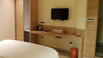 A hotel room with a bed, a TV mounted on the wall, a small table with a kettle, and a trash bin.