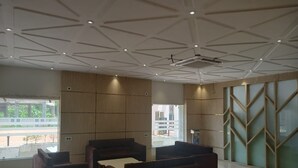 Um lobby espaçoso com móveis modernos, teto alto com padrões geométricos e grandes janelas com vista para o exterior.