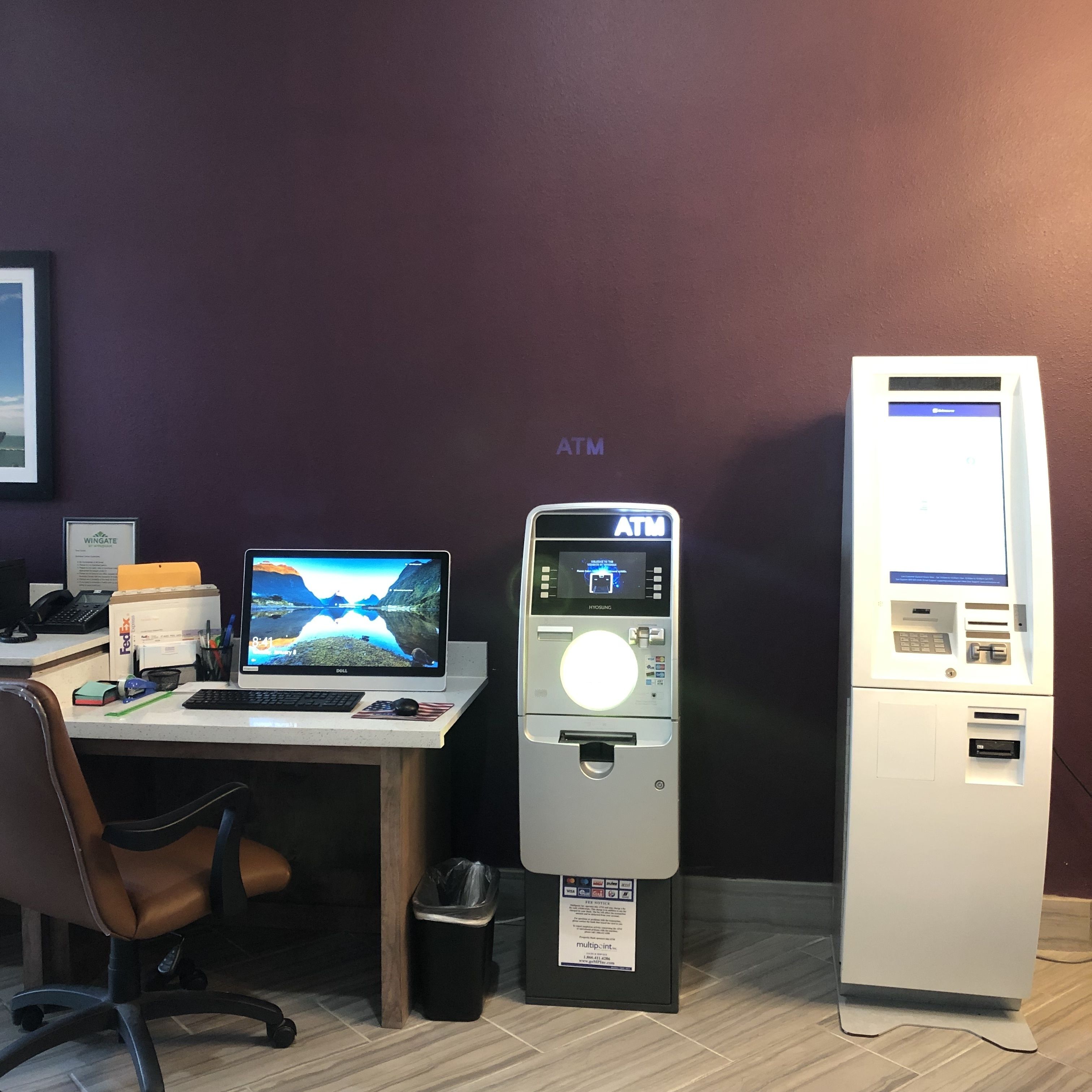 A desk with a computer, an ATM, and a trash can in a room with a purple wall.