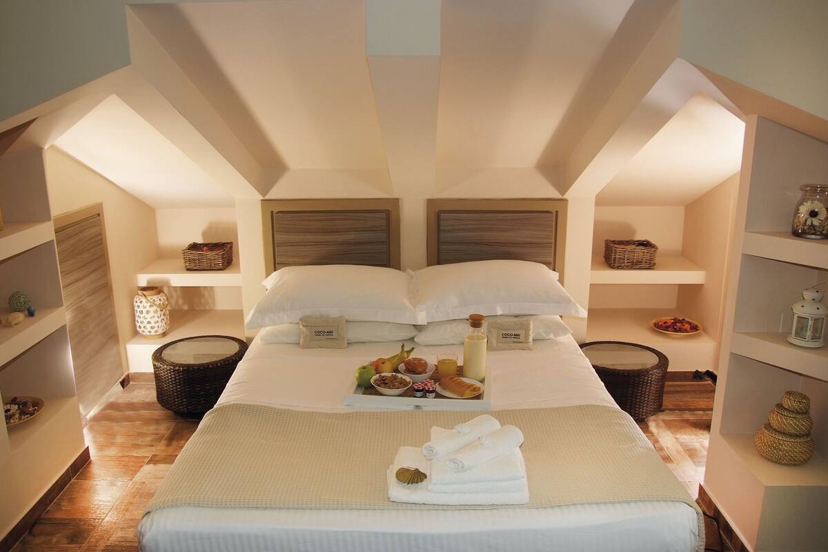 A hotel room with a large bed, two bedside tables, a basket, a bowl of fruit, and a bottle of milk.
