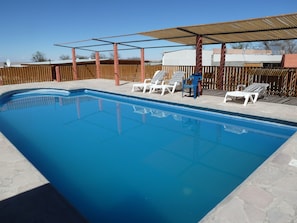 A swimming pool with clear blue water, surrounded by a wooden fence and a covered patio area with white plastic chairs.