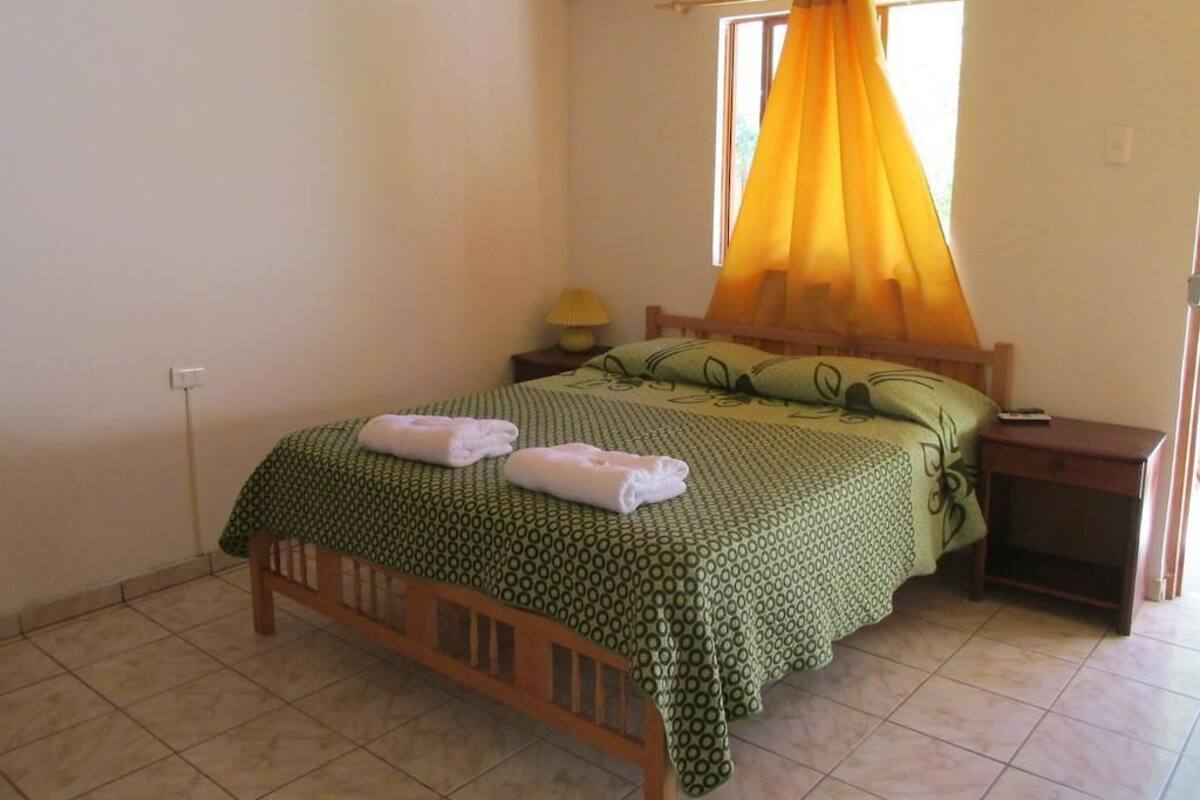 Uma cama de solteiro com colcha verde estampada, cabeceira de madeira e cortina amarela. Tem uma mesa de cabeceira com luminária e uma pequena prateleira.