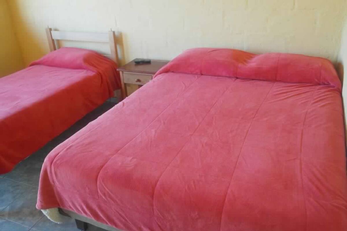 Quarto com duas camas de solteiro, cada uma com colcha e almofadas vermelhas. Um criado-mudo de madeira entre as camas tem um controle remoto sobre ele. As paredes são simples e o chão é revestido de azulejos.