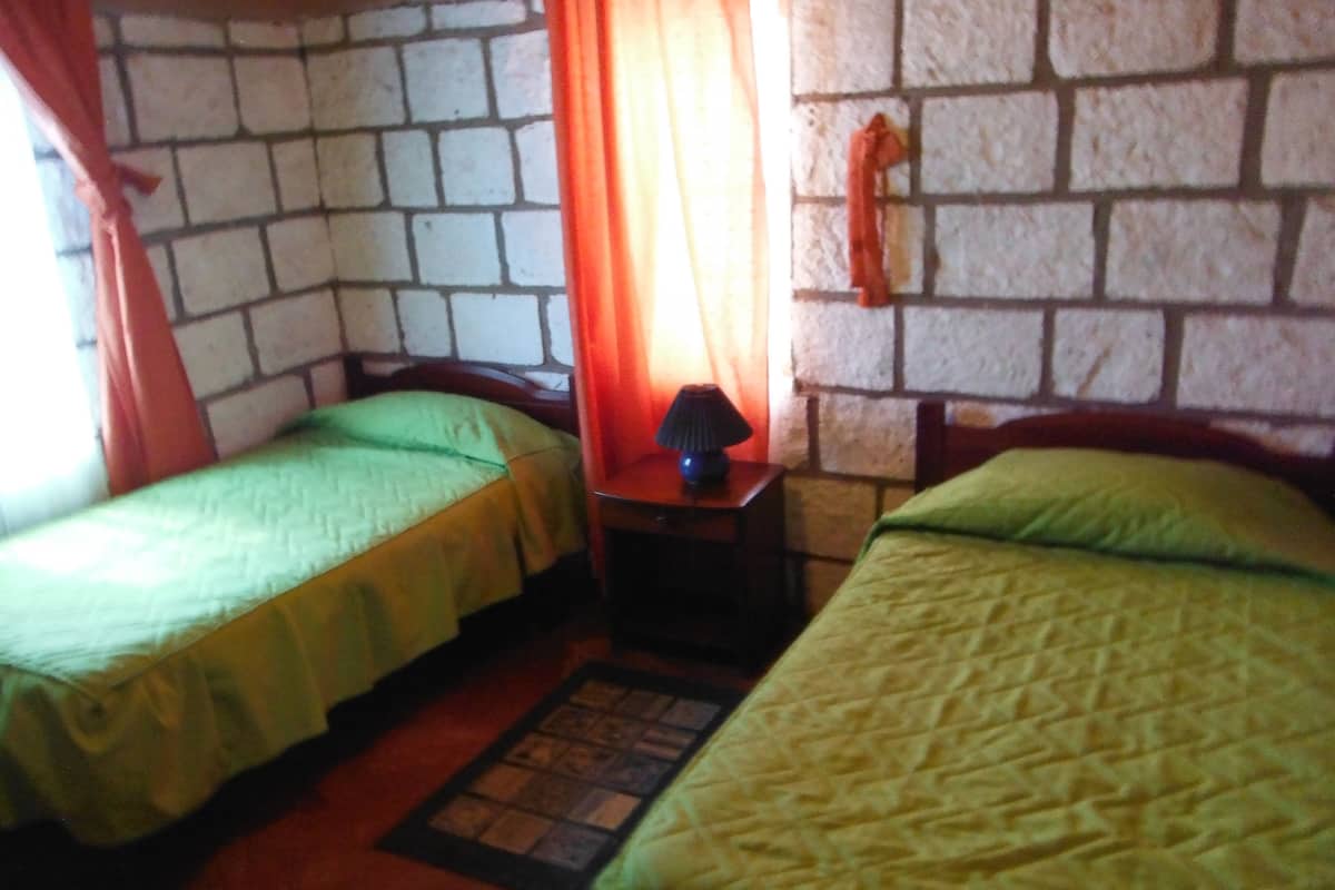 Quarto com duas camas de solteiro, uma mesinha com abajur e parede de pedra.