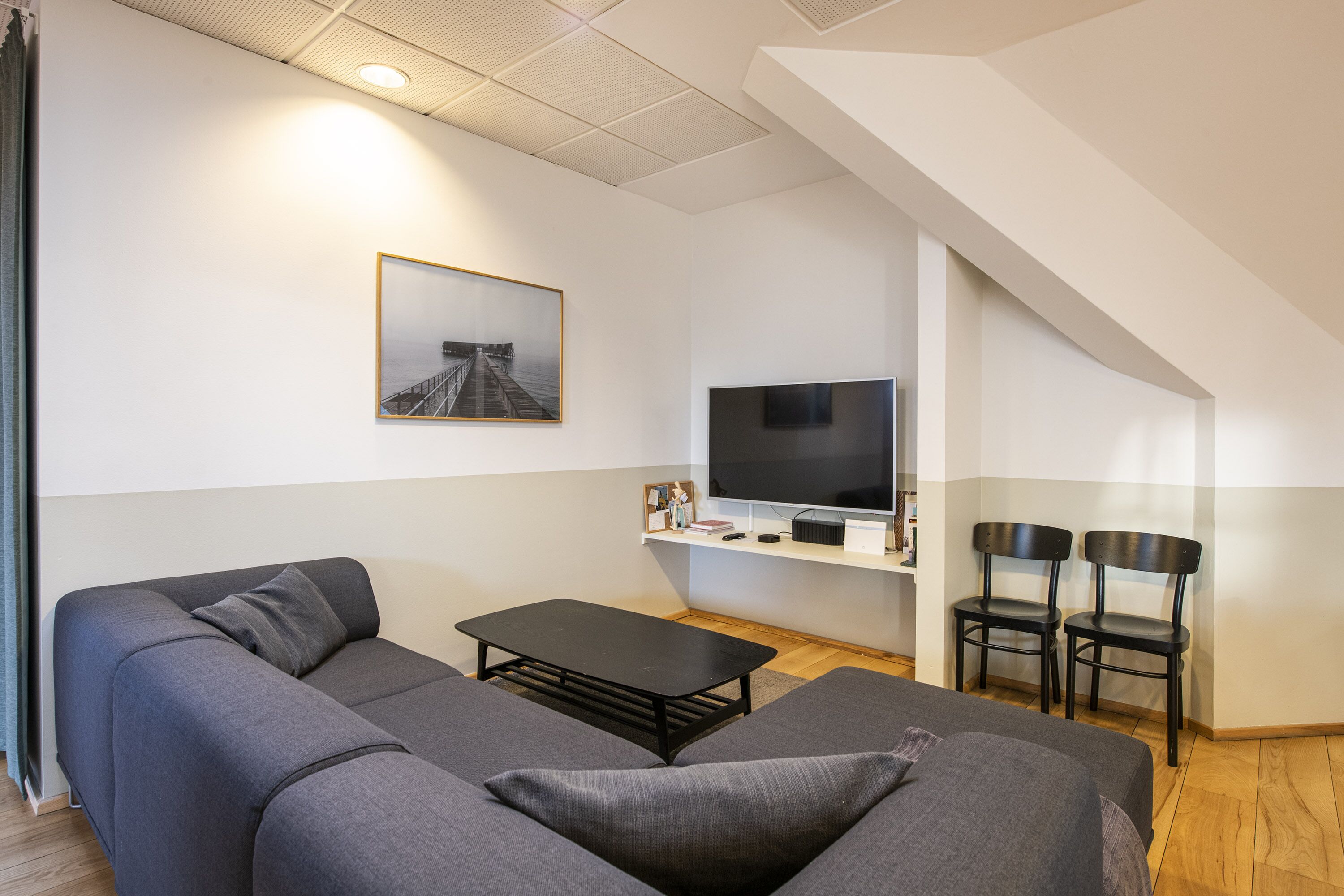 Ett modernt vardagsrum med en grå soffa, ett svart soffbord, en platt-TV på en vit hylla och två svarta stolar.