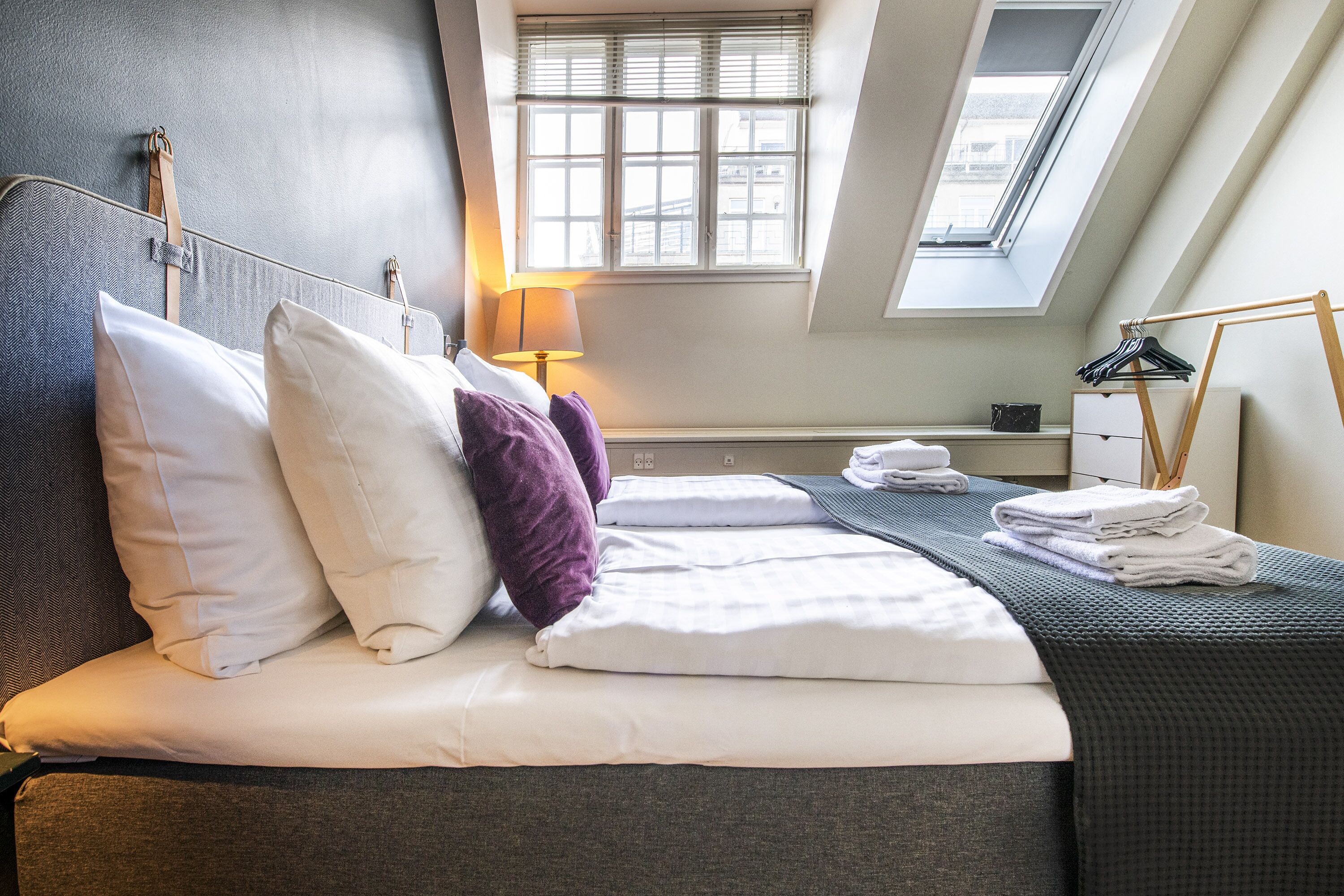 A neatly made bed with pillows and a blanket, a bedside table with a lamp, and a window with blinds.