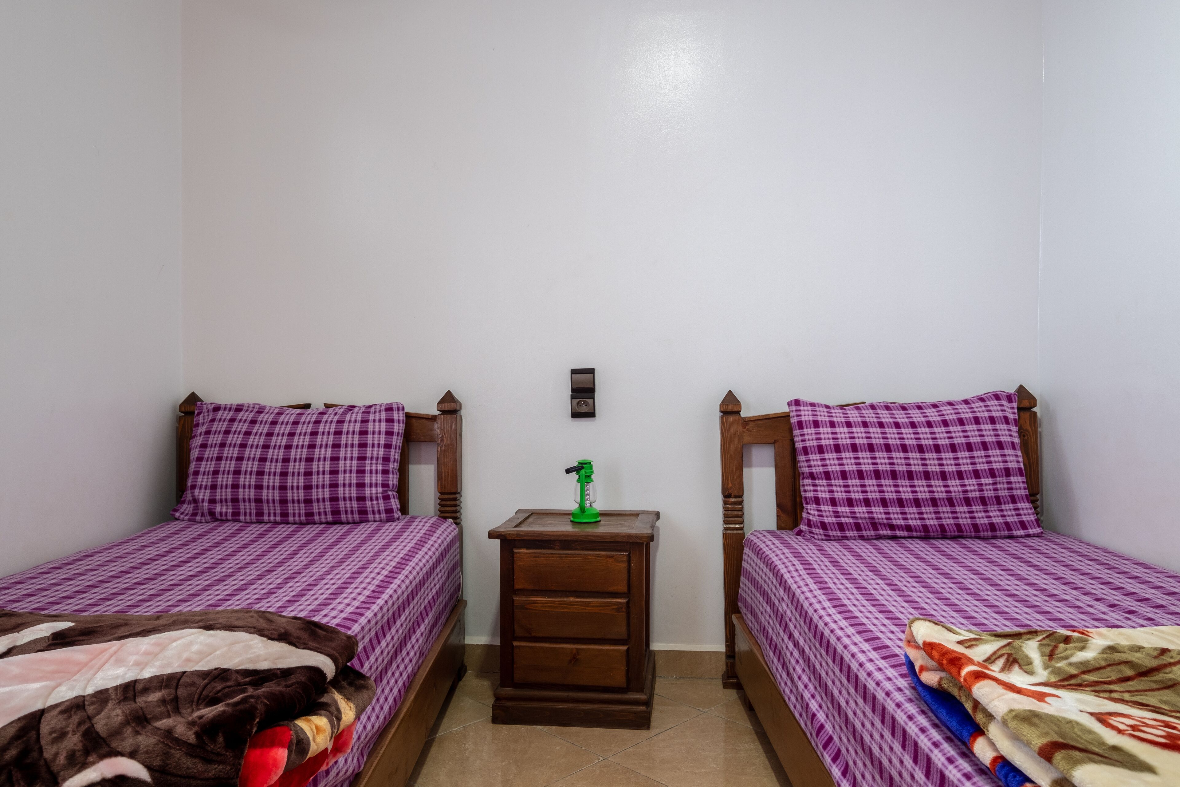 A small, clean room with two beds, each with purple bedding, a wooden nightstand with a green object, and a small picture on the wall.