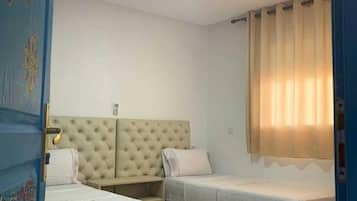 Une chambre d’hôtel avec deux lits, une tête de lit capitonnée, une fenêtre avec des rideaux et une porte ornée de panneaux décoratifs.