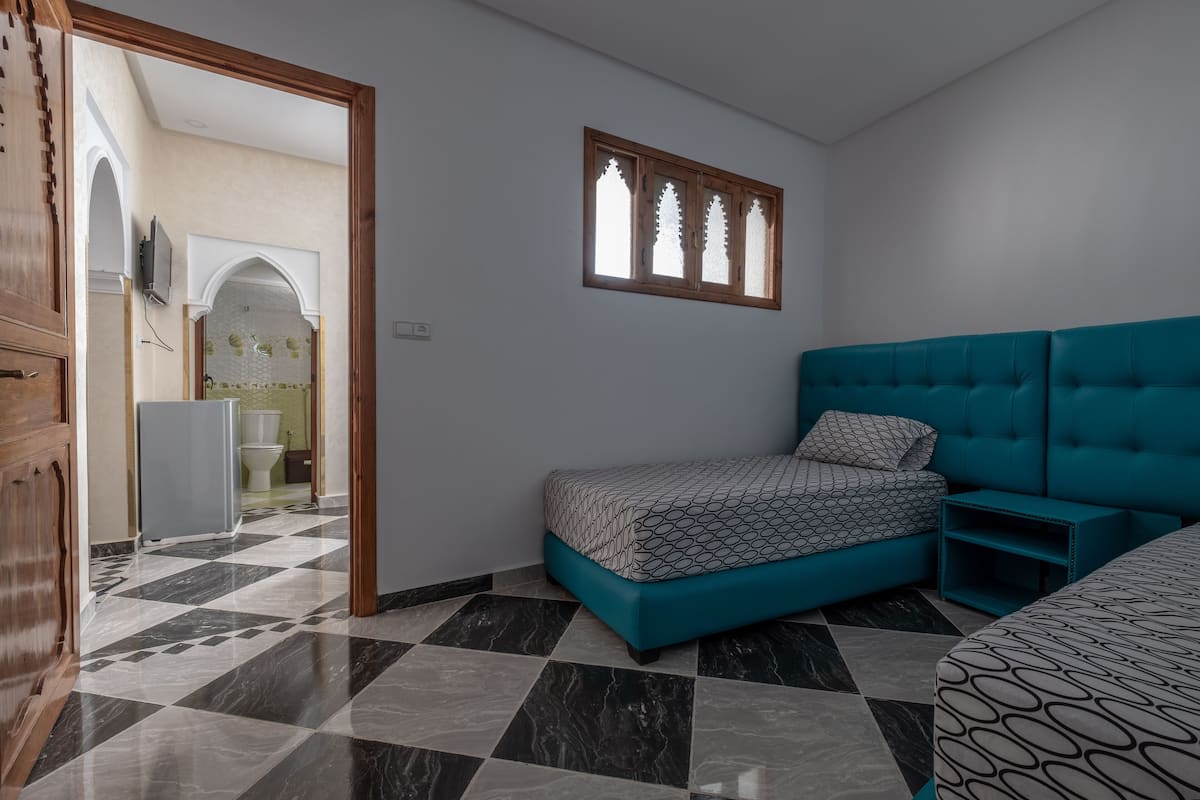 A bedroom with a checkered floor, a teal sofa bed, a patterned bedspread, and a window with a wooden frame.