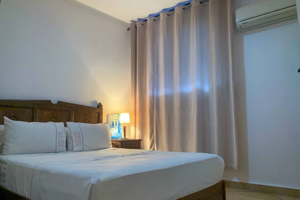 A hotel room with a bed, curtains, an air conditioning unit, and a nightstand with a lamp.