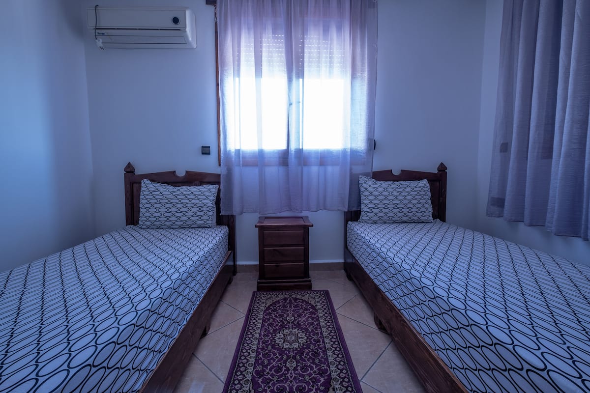 A room with two beds, a small wooden nightstand, a patterned rug, and a window with sheer curtains.