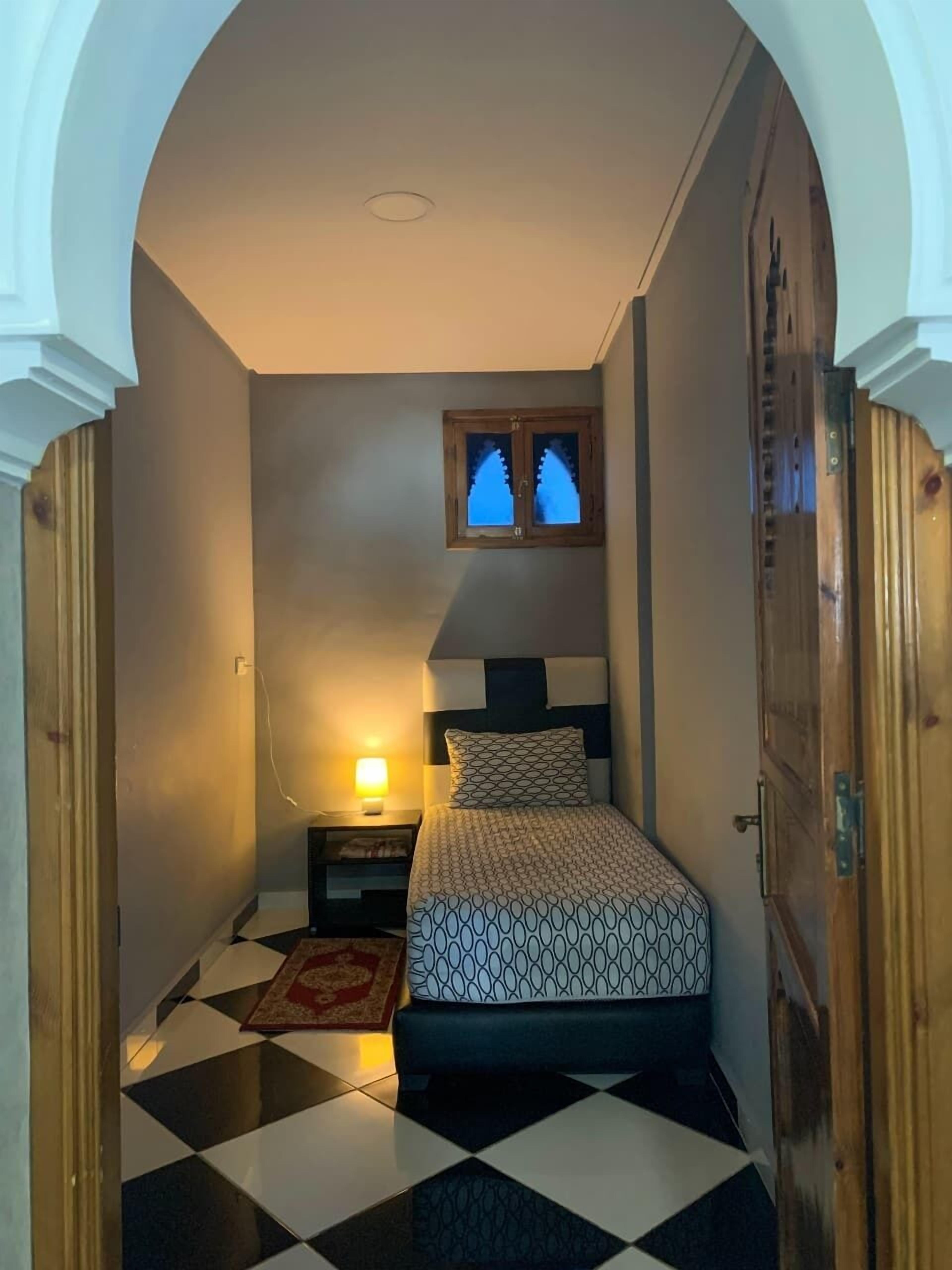 A bedroom with a bed, a nightstand, a lamp, a window, and a checkered floor.
