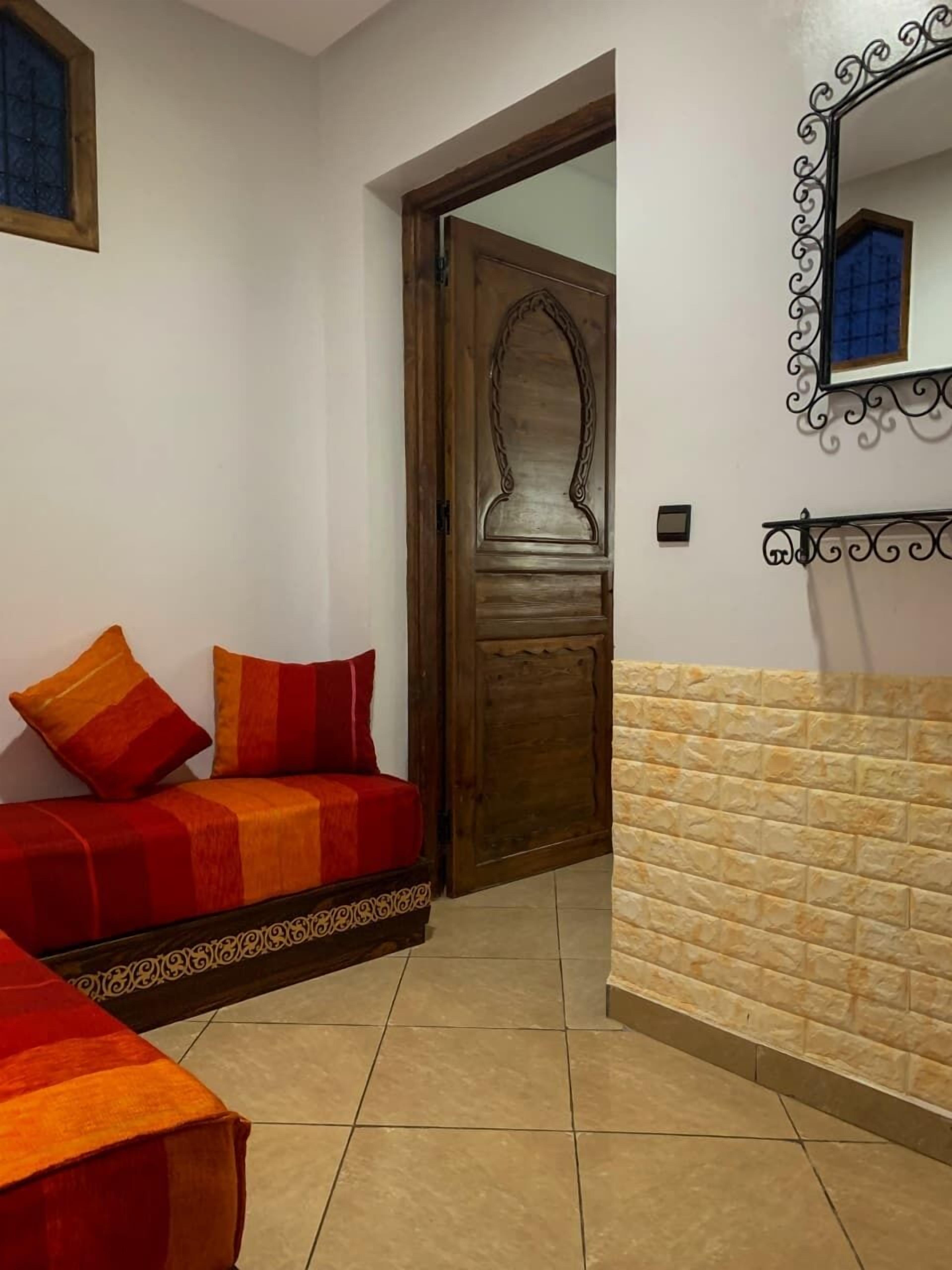 A room with a wooden door, a mirror with a decorative frame, a red sofa with cushions, and a tiled floor.