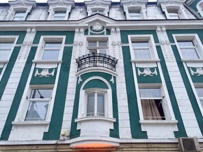Un edificio con fachada verde y blanca, detalles arquitectónicos ornamentales y un balcón con barandilla de hierro forjado.