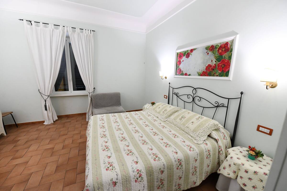Una camera da letto con un letto, una sedia, un tavolino, un quadro e una finestra con tende.